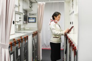 A woman in a flight attendant’s uniform taps on a tablet in an airplane galley.