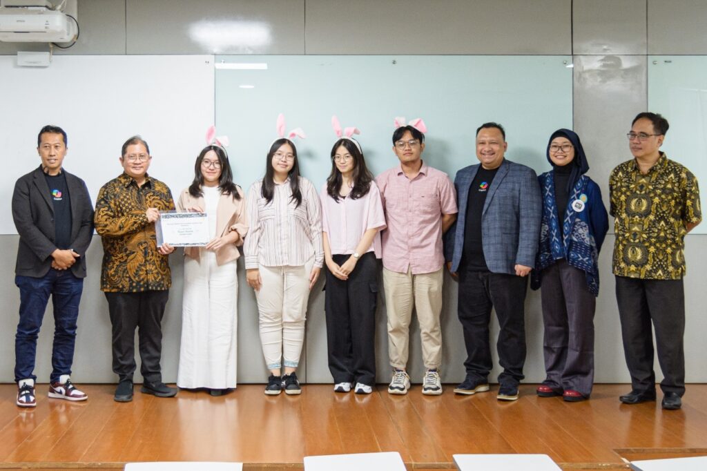 Eleven people standing in a row indoors. Five young people in the center wear pink bunny ear headbands, with one holding a certificate. The group includes men and women in formal and casual clothing, posing in front of a whiteboard and glass panel in a classroom or office setting.