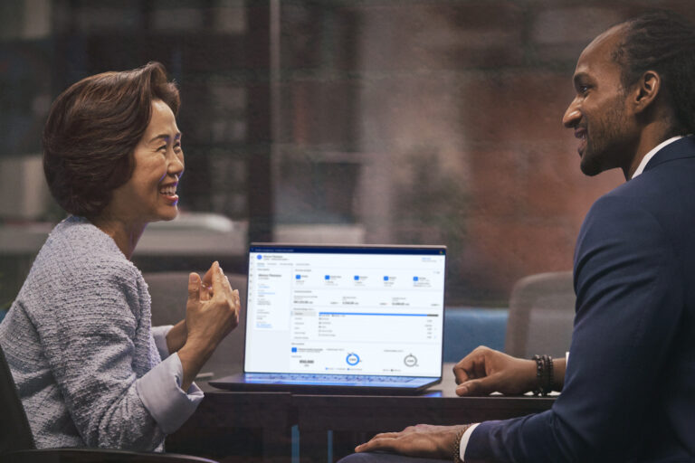 Two people sitting at a table in an office, viewing a laptop screen displaying a financial dashboard with account balances, recent transactions, pie charts, and bar graphs. One person is gesturing while the other looks attentively at the screen.